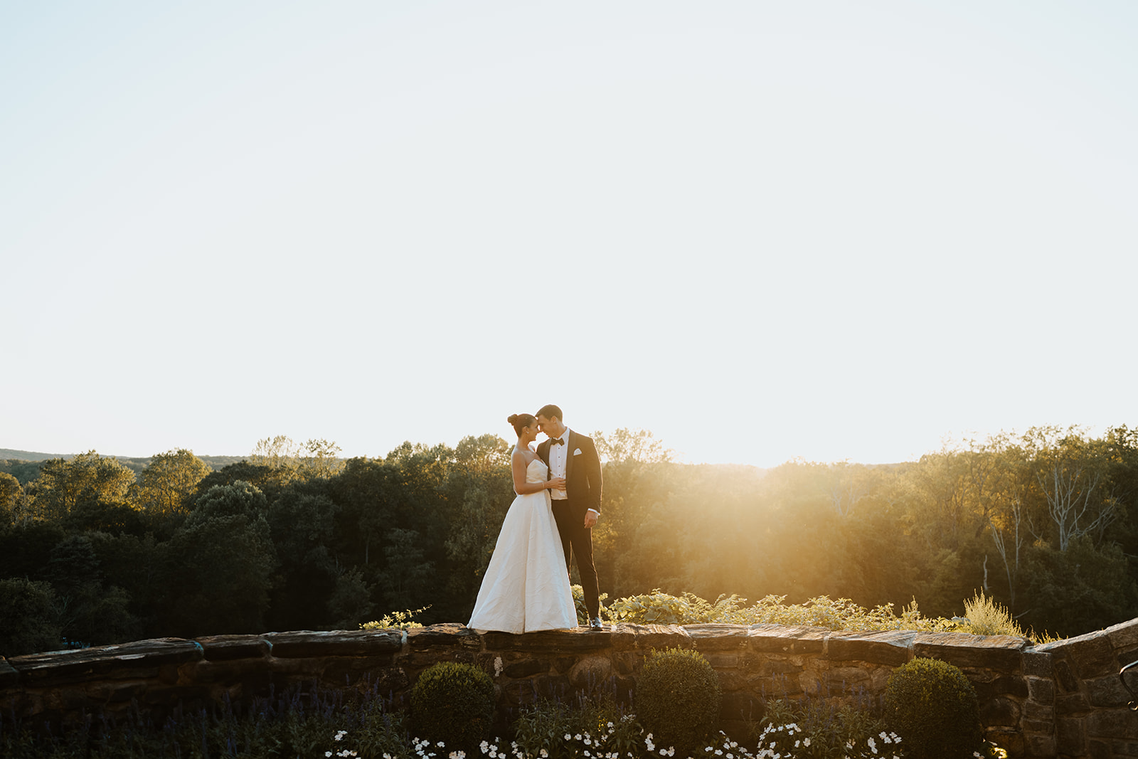 Newlyweds standing on a stone wall at sunset, touching foreheads against a bright golden sky.